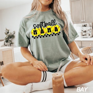 Op de afbeelding: Een vrouw draagt een lichtgroen T-shirt met de tekst "Softball Mama" en een afbeelding van vijf gele softbal met zwarte naden. Het shirt wordt gecombineerd met een witte broek en witte sokken met zwarte strepen.