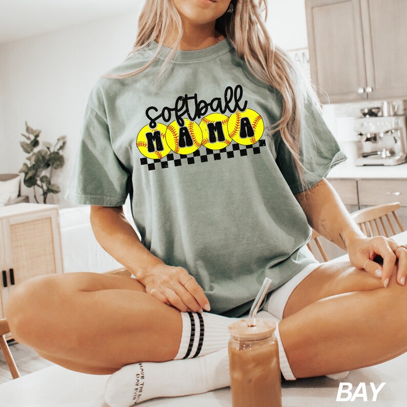 Op de afbeelding: Een vrouw draagt een lichtgroen T-shirt met de tekst "Softball Mama" en een afbeelding van vijf gele softbal met zwarte naden. Het shirt wordt gecombineerd met een witte broek en witte sokken met zwarte strepen.