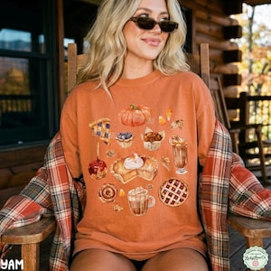 May include: A burnt orange crewneck t-shirt featuring a graphic print of various autumn-themed treats including a pumpkin, a slice of pie, a caramel apple, a cinnamon roll, a latte, and a muffin, layered under a red and orange plaid flannel shirt.