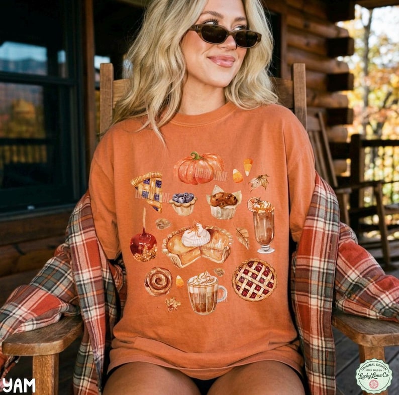 May include: A burnt orange crewneck t-shirt featuring a graphic print of various autumn-themed treats including a pumpkin, a slice of pie, a caramel apple, a cinnamon roll, a latte, and a muffin, layered under a red and orange plaid flannel shirt.