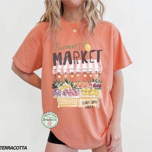 May include: Terracotta-colored t-shirt with a Farmers Market graphic. The design features a market stall with fruits and vegetables, the text "Farmers Market", and the hours "10AM-5PM DAILY". The shirt has short sleeves.