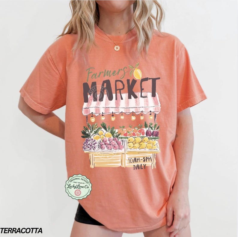 May include: Terracotta-colored t-shirt with a Farmers Market graphic. The design features a market stall with fruits and vegetables, the text "Farmers Market", and the hours "10AM-5PM DAILY". The shirt has short sleeves.