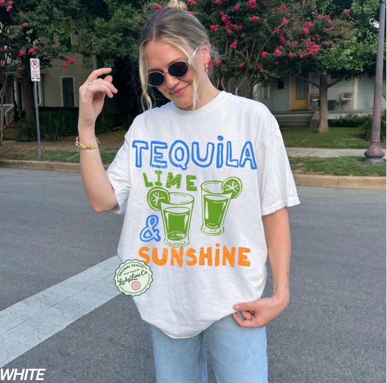 May include: White t-shirt featuring the text "TEQUILA LIME & SUNSHINE" in blue, green, and orange. The design includes two green cocktail glasses with lime slices. The shirt has a relaxed, oversized fit.
