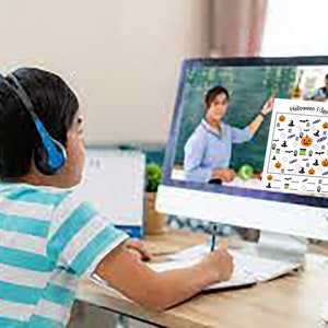 May include: A child wearing blue headphones sits at a desk in front of a computer screen. The screen shows a teacher on a video call and a Halloween I Spy printable activity sheet.