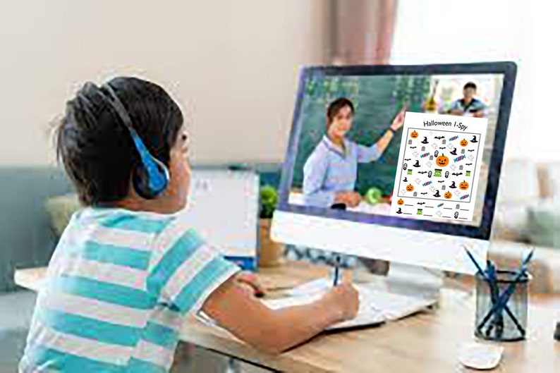 May include: A child wearing blue headphones sits at a desk in front of a computer screen. The screen shows a teacher on a video call and a Halloween I Spy printable activity sheet.