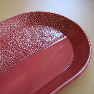 May include: A red ceramic serving tray with a floral embossed design on one half and a smooth surface on the other. The tray has an oval shape and is suitable for serving food or displaying decorative items.
