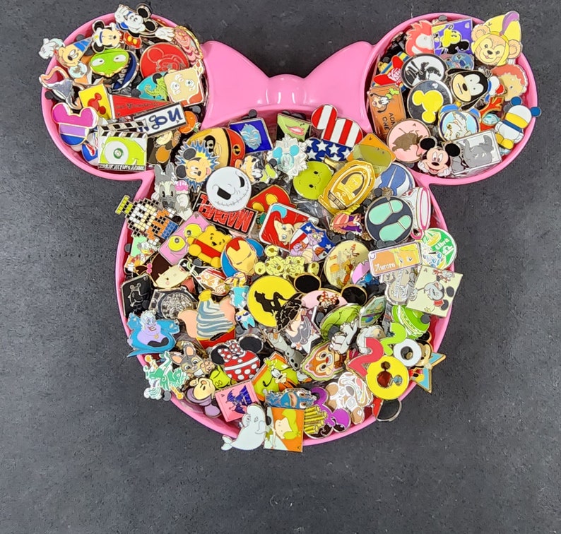 May include: A pink Mickey Mouse shaped container filled with a variety of colorful Disney character pins. The pins feature characters such as Mickey Mouse, Minnie Mouse, Donald Duck, Goofy, Pluto, and many others. The pins are arranged in a random pattern and some of the pins have glitter or other embellishments.