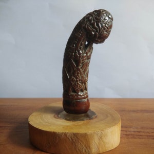 Op de afbeelding: Een gegraveerd houten handvat met ingewikkelde details, gemonteerd op een houten basis. Het handvat is donkerbruin en heeft een gebogen vorm.