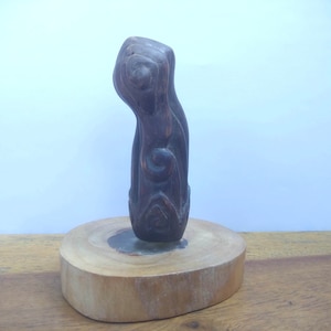 Puede incluir: Una escultura de madera marrón oscuro de una persona con la cabeza inclinada. La escultura está de pie sobre una base redonda de madera.