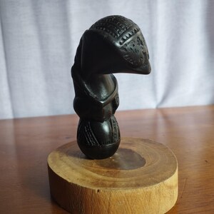 Può includere: Una scultura in legno nero di una persona con la testa rotonda e un design intagliato. La scultura è in piedi su una base di legno.
