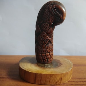 May include: A carved wooden handle with intricate details, featuring a bird design. The handle is mounted on a wooden base.