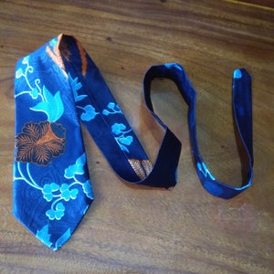 Op de afbeelding: Een blauwe zijden stropdas met een bloemenpatroon in tinten oranje, blauw en wit. De stropdas heeft een donkerblauwe voering.