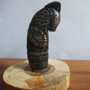May include: A dark brown and green carved wooden handle with intricate details. The handle is shaped like a bird's head and has a textured surface.