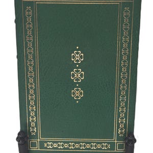 May include: A green leather-bound book with gold accents and a decorative border. The book is on a black metal stand.