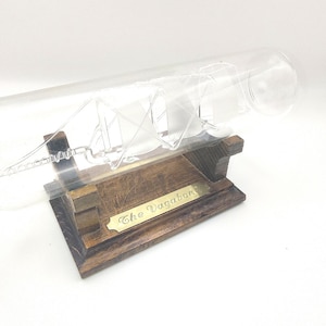 May include: A clear glass ship model inside a glass tube, mounted on a dark wooden base. The base has a brass plate with the inscription "The Vagabond". The ship's rigging is visible inside the tube.