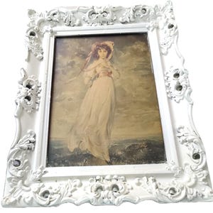 May include: A vintage framed print featuring a woman in a flowing gown and hat. The print is set within a white, ornate, baroque-style frame. The frame has detailed floral and scrollwork designs, adding a classic touch to the artwork.