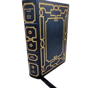 May include: A hardcover book titled "Beggerman, Thief" by Irwin Shaw. The book has a dark blue cover with gold decorative accents and text. The spine features the title, author, and "First Edition" in gold lettering. A black ribbon bookmark is visible.