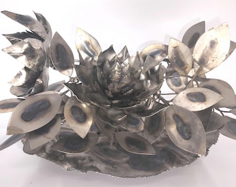 May include: A silver metal sculpture of a flower with multiple leaves and petals. The sculpture is made of welded metal and has a rustic, industrial look.