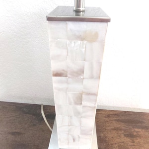 May include: A decorative table lamp with a square base and a rectangular body. The body is covered in a mosaic of white and tan squares. The lamp has a silver metal base and a silver metal top. The lamp is turned on and has a white light.