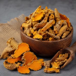 Puede incluir: Un cuenco de madera lleno de raíces de cúrcuma secas, con un interior naranja vibrante y un exterior marrón texturizado. Varios trozos de cúrcuma cortados están esparcidos cerca, acompañados por una cuchara de madera con más segmentos.