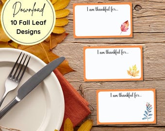 May include: A wooden table setting with fall-themed elements. Includes white place settings, a fork, knife, and three orange-bordered cards with the text "I am thankful for...". The cards feature watercolor leaf illustrations.