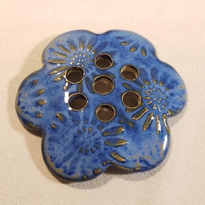 May include: A blue ceramic flower frog with six holes for arranging flowers. The flower frog has a textured surface with gold accents and a flower-like design. The flower frog is a decorative item for floral arrangements.