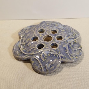 May include: A ceramic flower frog in a light blue hue with yellow speckles. The frog has a floral design with seven holes for arranging flowers. The edges are shaped like a flower, adding to its decorative appeal.