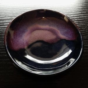 May include: A small, round ceramic bowl with a glossy finish. The bowl's interior features a blend of dark purple, burgundy, and black hues, creating a marbled effect. The rim is smooth and reflective, and the bowl is set against a dark background.