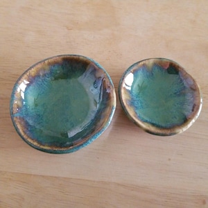 May include: Two handmade ceramic bowls with a glossy, teal glaze and a brown and gold rim. The bowls are different sizes and have an organic, irregular shape. The bowls are likely used for serving small portions of food or as decorative items.