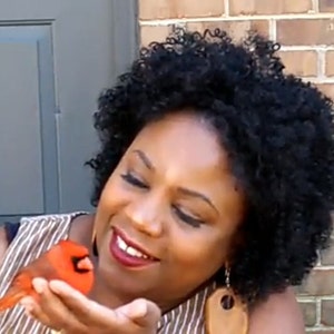 May include: A woman with curly black hair smiles as she holds a red cardinal in her hand.