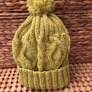 Könnte beinhalten: Eine grüne Strickmütze mit Zopfmuster und einem flauschigen Bommel oben.