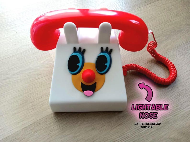 Op de afbeelding: Een witte speelgoedtelefoon met een rode draaischijf en een cartoon gezicht. Het gezicht heeft grote blauwe ogen, een gele mond en een rode neus die oplicht. De tekst "LIGHTABLE NOSE" en "BATTERIES NEEDED - TRIPLE A" staat onder de telefoon.