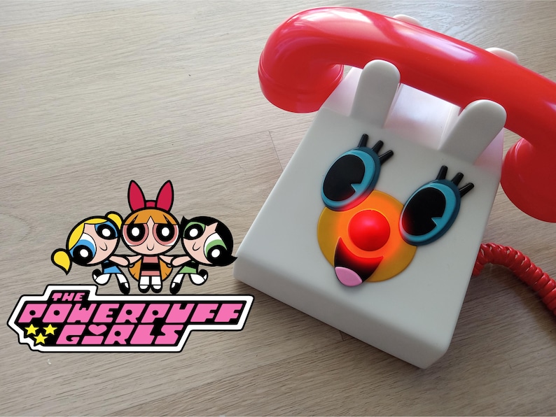 Op de afbeelding: Een witte speelgoedtelefoon met een rode draaischijf en een glimlachend gezicht. De telefoon heeft een rode knop op het gezicht en blauwe ogen. Het Powerpuff Girls-logo staat op de voorgrond.