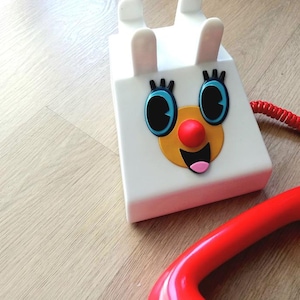 Op de afbeelding: Een witte speelgoedtelefoon met een rood snoer en een rode hoorn. De telefoon heeft een cartoon gezicht met blauwe ogen, een gele neus en een rode mond.