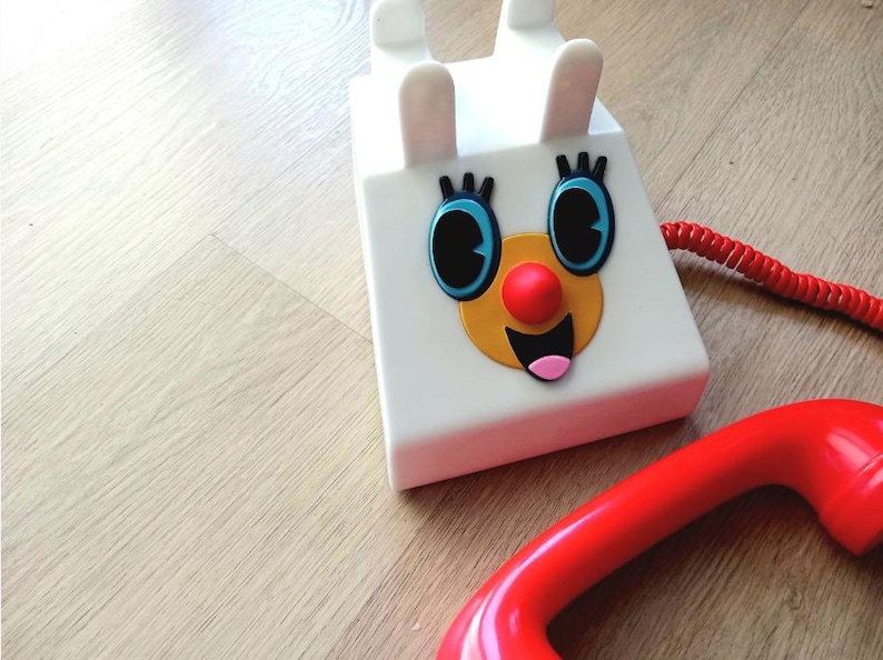 Op de afbeelding: Een witte speelgoedtelefoon met een rood snoer en een rode hoorn. De telefoon heeft een cartoon gezicht met blauwe ogen, een gele neus en een rode mond.