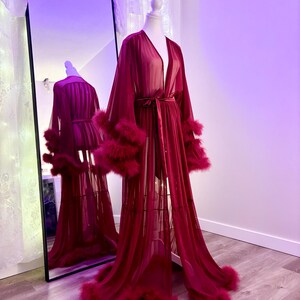 Puede incluir: Una larga bata roja transparente con ribete de plumas alrededor de las mangas y el dobladillo. La bata tiene un cintur&oacute;n de lazo en la cintura.