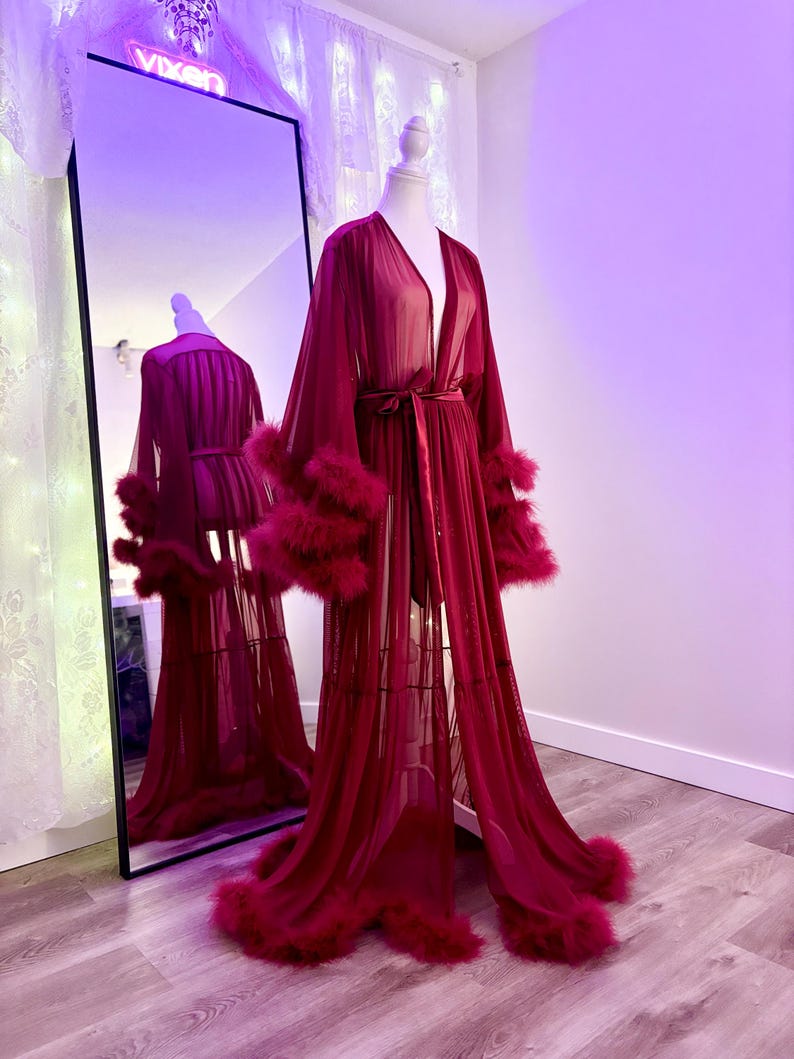 Puede incluir: Una larga bata roja transparente con ribete de plumas alrededor de las mangas y el dobladillo. La bata tiene un cintur&oacute;n de lazo en la cintura.