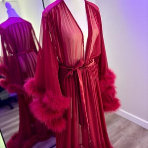 Puede incluir: Una larga bata roja transparente con ribetes de plumas en las mangas. La bata tiene un escote en V y un cintur&oacute;n de lazo en la cintura.