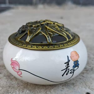May include: White ceramic incense burner with a bronze-colored lid. The burner features a pink lotus flower and a cartoon monk with Chinese characters. The lid has an intricate, openwork design.