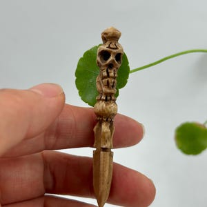May include: A carved wooden dagger featuring a skull design at the top. The dagger is light brown in color and has intricate details. The skull has visible eye sockets and a detailed jawline. The dagger is held in a hand against a white background.