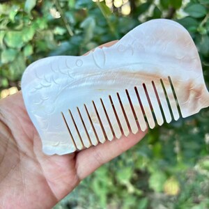 May include: A pearlescent, carved comb with a decorative design. The comb features a series of evenly spaced teeth and a curved, ornate handle. The color is primarily white with hints of tan and brown.