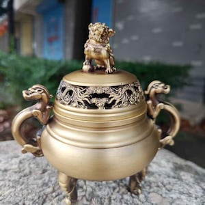 Puede incluir: Un quemador de incienso dorado con una tapa decorativa y asas en forma de dragón. Una pequeña estatua de león se encuentra sobre la tapa. El quemador tiene tres patas ornamentadas y está colocado sobre una superficie de piedra texturizada.