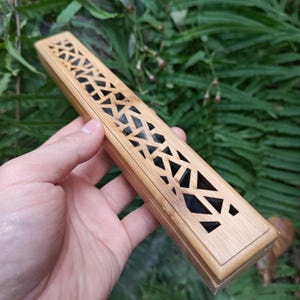 May include: A rectangular, light brown wooden incense holder with a geometric cut-out design on the lid. The holder is held in a hand, with a green leafy background.