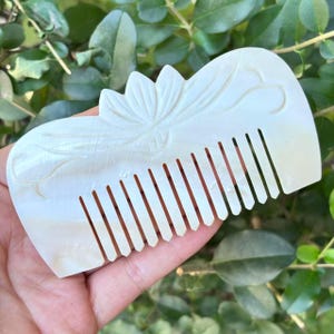 May include: A pearlescent white comb with a carved floral design. The comb features a flower at the top and intricate leaf and vine details. The teeth are evenly spaced and the comb is held against a background of green leaves.