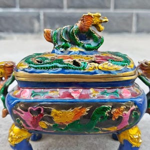 Puede incluir: Un quemador de incienso ornamentado con un motivo de dragón. La pieza de cerámica presenta una base azul, detalles dorados y un dragón multicolor en la parte superior. Los lados están decorados con dragones y motivos florales.