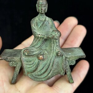 May include: A bronze-colored figurine of a seated figure on a bench. The figure is wearing a robe and holding a cloth. The bench has a simple design with four legs. The figurine has a weathered appearance with green and brown tones.