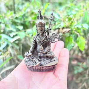 May include: A bronze-colored statue of a seated figure, holding a staff and a small object. The figure is adorned with detailed clothing and a headdress. The statue is resting on a decorative base, with a blurred green background.