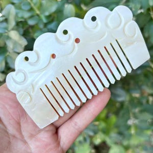 May include: A white, carved comb with a cloud-like design along the top edge. The comb features evenly spaced teeth and small circular accents. The comb is held in a hand against a blurred green background.