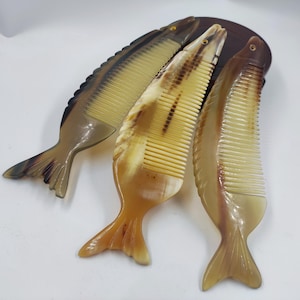 May include: Three fish-shaped combs in shades of brown and tan. The combs have a fish tail design and a comb on the back. The combs are made of a light-colored material.