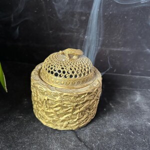 May include: A gold-colored incense burner with a detailed, textured design. The lid features a honeycomb pattern and a small frog figurine. Smoke rises from the burner, suggesting it is in use. The base has a carved leaf pattern.